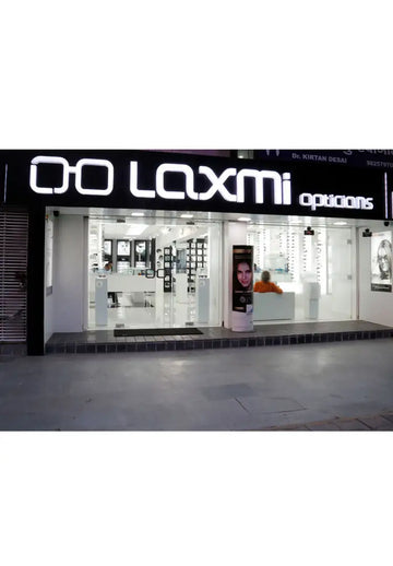 The illuminated black sign for ’laxmi opticians’ with white glowing lettering and a distinctive logo.