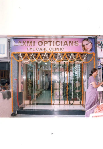 The storefront sign for laxmi opticians eye care clinic features bold gold lettering on a purple background with an image of a woman wearing glasses.